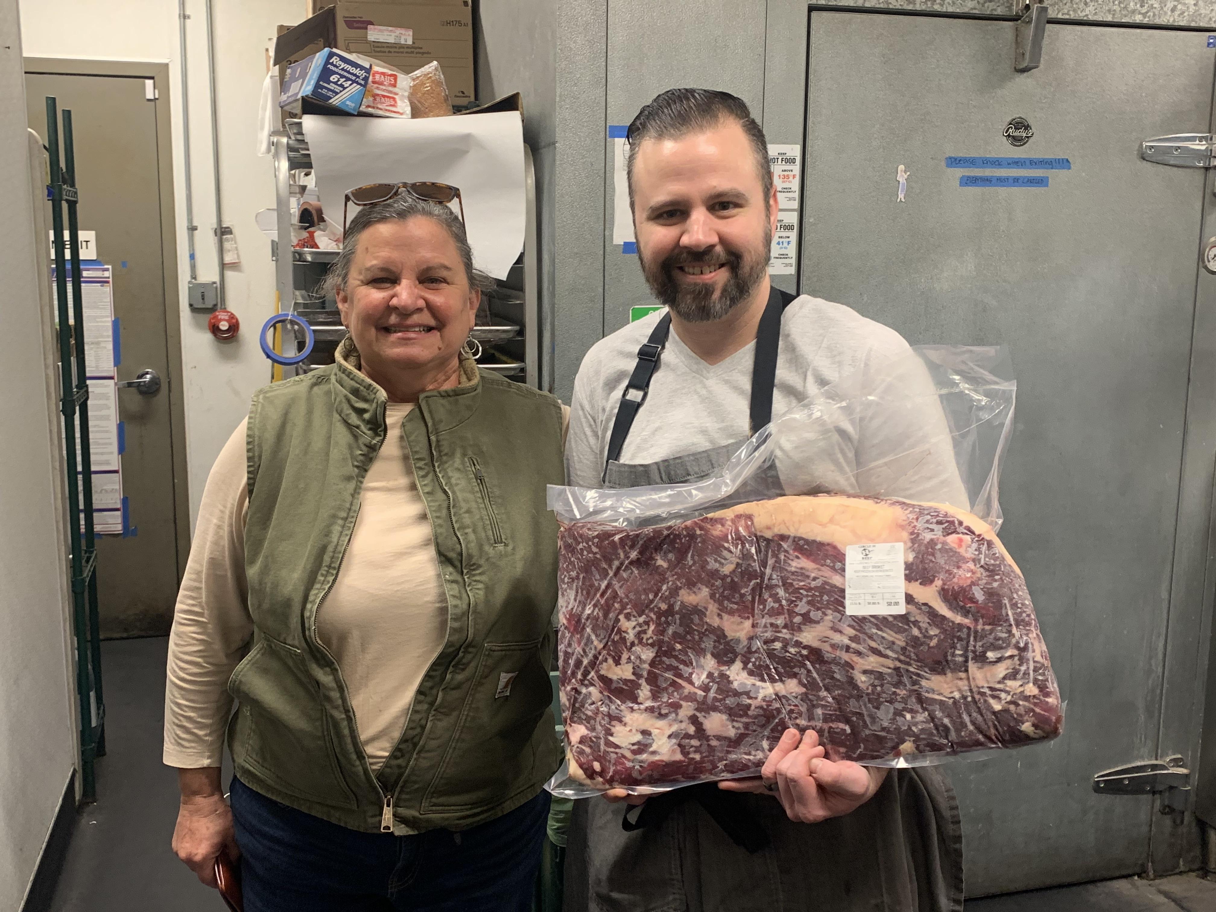 Woman wearing a vest with a man holding a cut of beef.