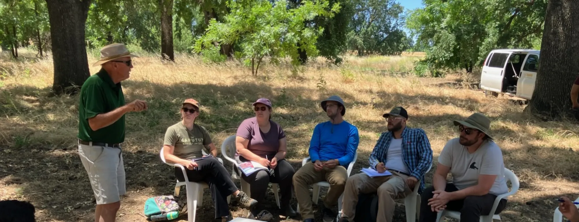 Man in polo and shorts and hat at left speaks with a group of students under shade trees during a farm tour