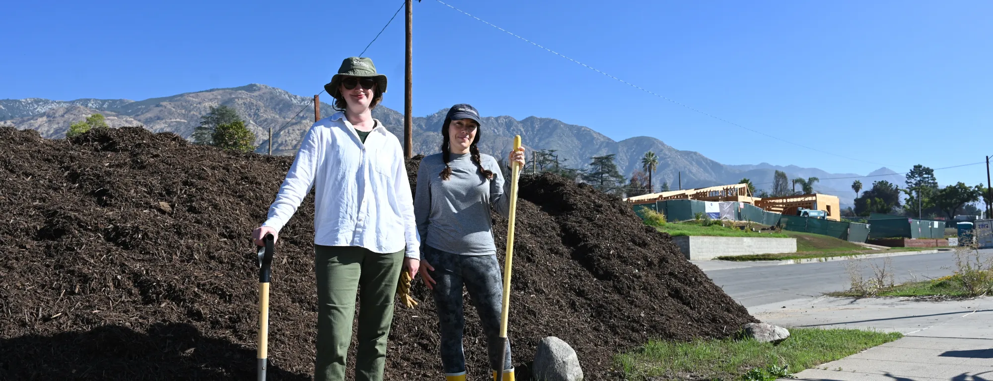 Two people with shovels stand in front of a pile of compost.
