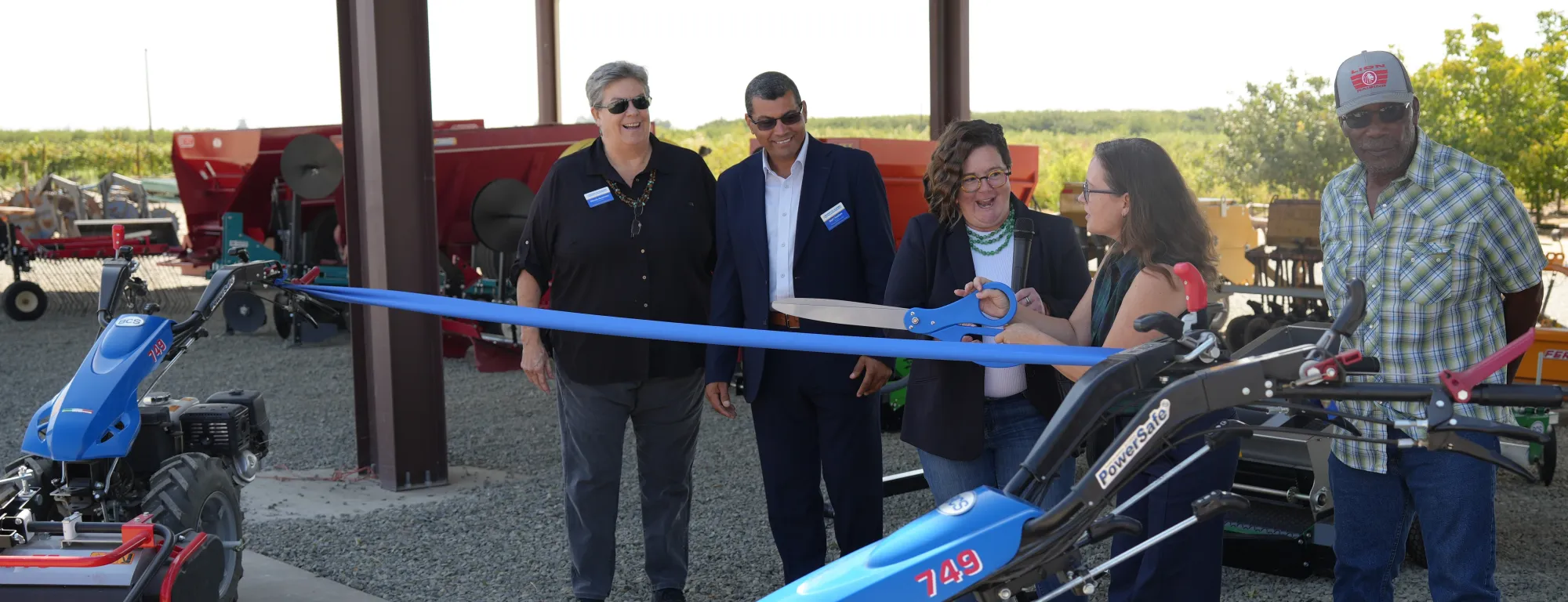 A group of people prepare to cut a ribbon strung between two pieces of farm equipment