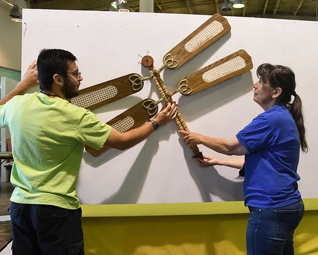 Preparing to hang a dragonfly project are McCormack Hall superintendent Gloria Gonzalez and assistant Daniel Brown, both of Vallejo. Crafted by Tina Saravia of Suisun City, it is made of recyclables: fan blades and furniture legs. (Photo by Kathy Keatley Garvey)