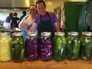 2 master food preservers smiling behind a row of fermented and pickled foods in glasses.