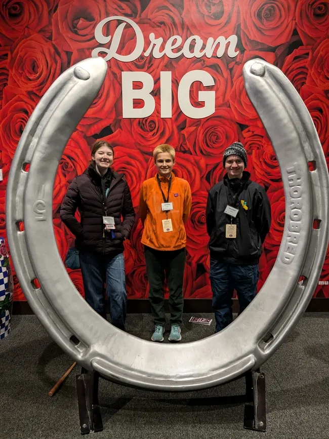 3 youth standing behind a large horseshoe sculpture,under