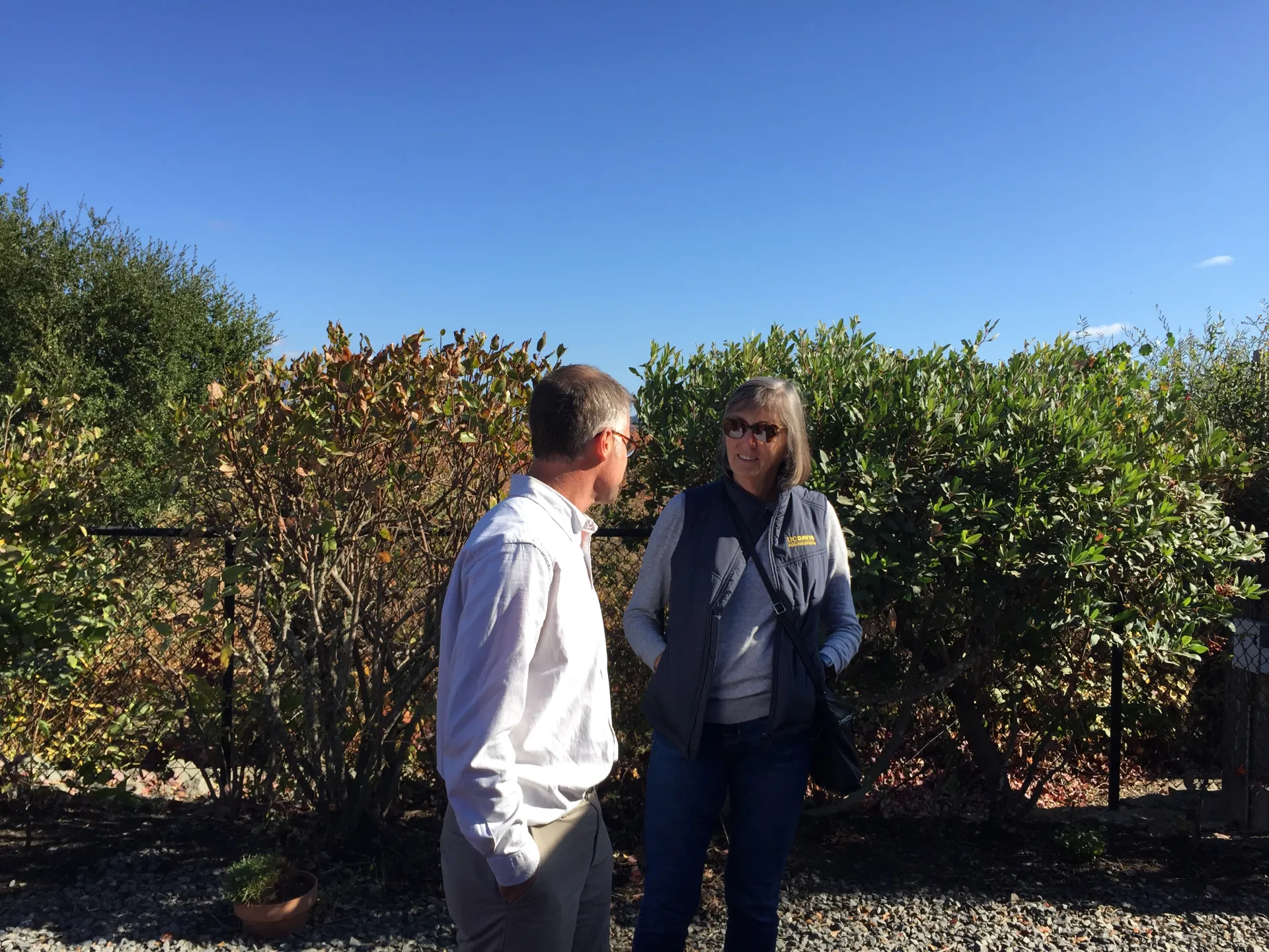 Swain and Stegura converse outdoors with shrubs in the background.