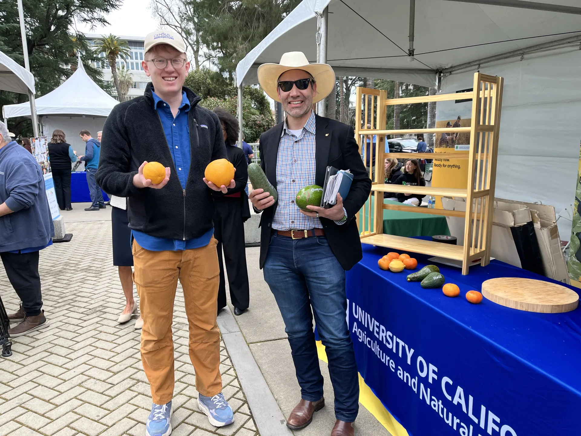 Jay, on left, holds 2 oranges, while Juan, wearing a cowboy hat and sunglasses, holds 2 large avocados. 