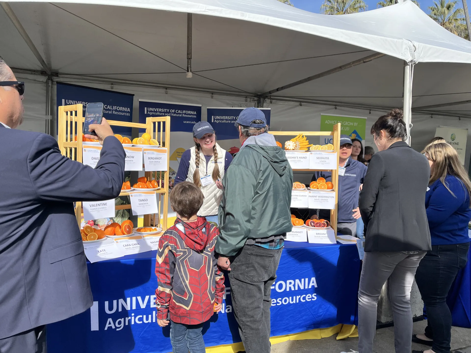 Judi, standing between shelves of citrus varieties, speaks with a man and a child while a man in a suit takes a photo.