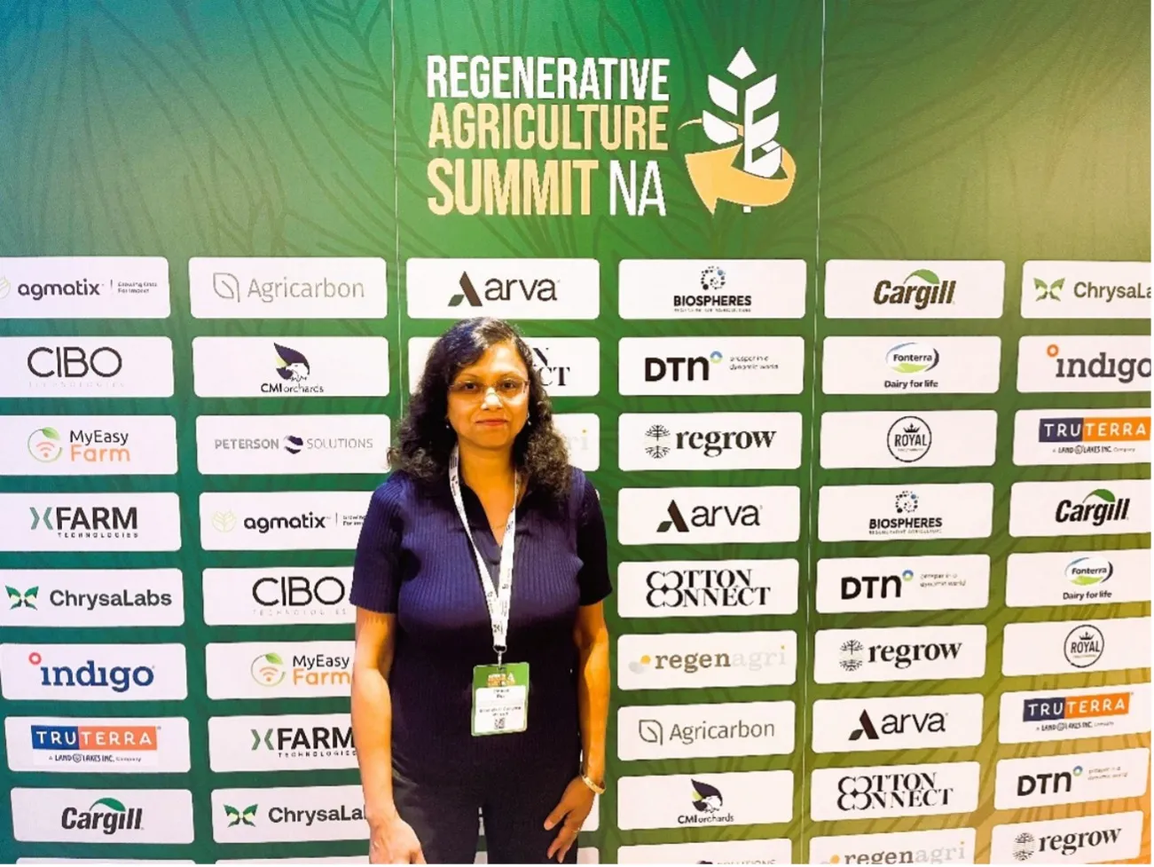 Srabani Das stands in front of a banner for Regenerative Agriculture Summit NA with sponsor logos. 