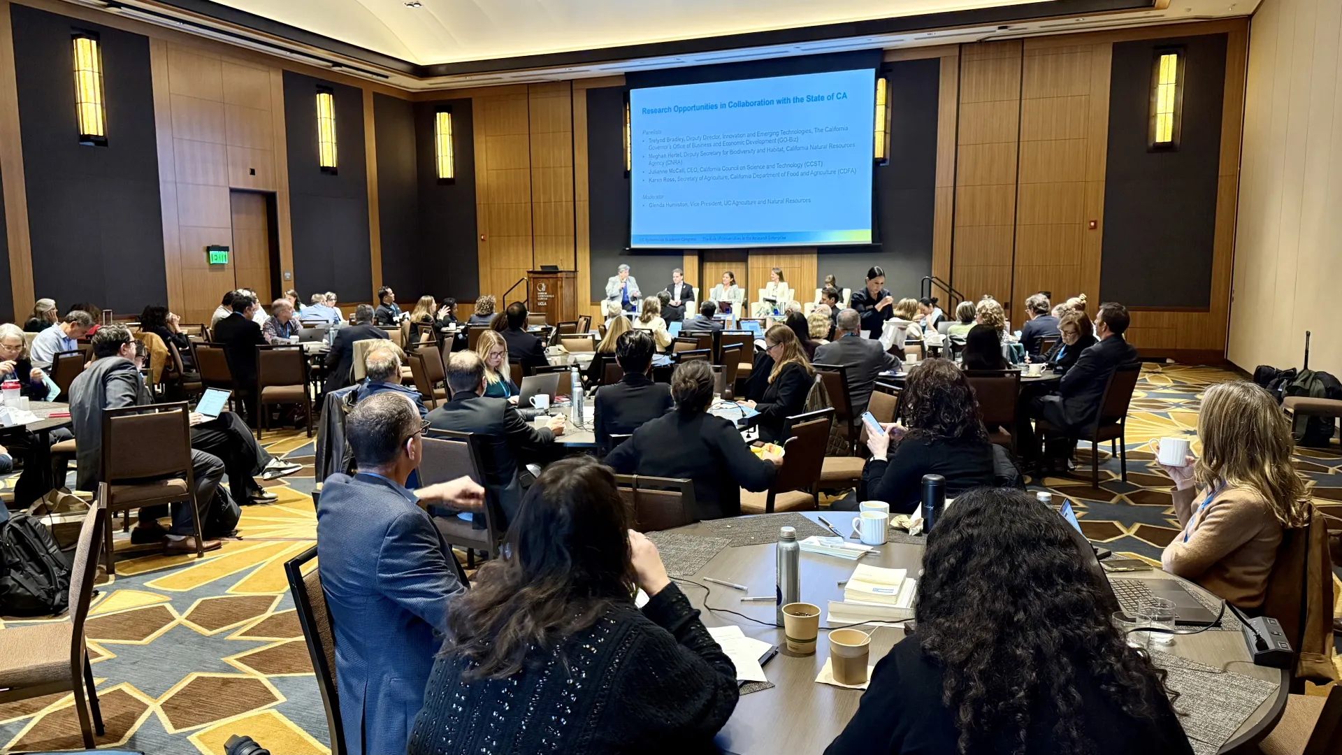 Glenda Humiston speaks while seated onstage with four panelists. Attendees sit at round tables.