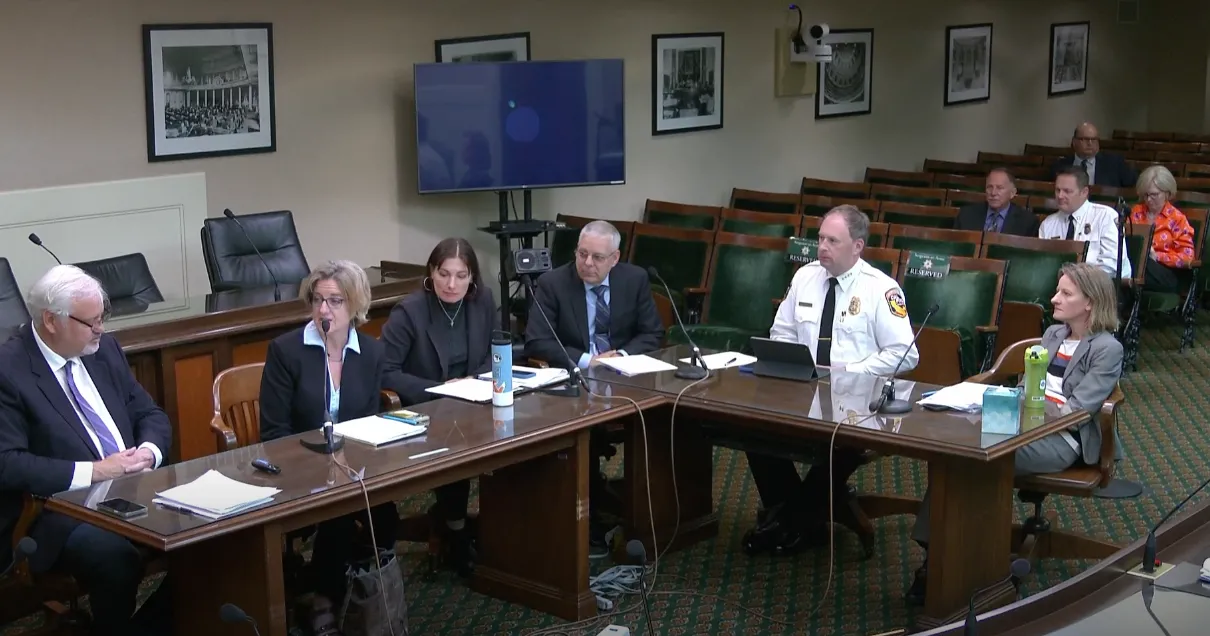 Yana speaks into a microphone as 6 other people seated with her at an L-shaped table listen.