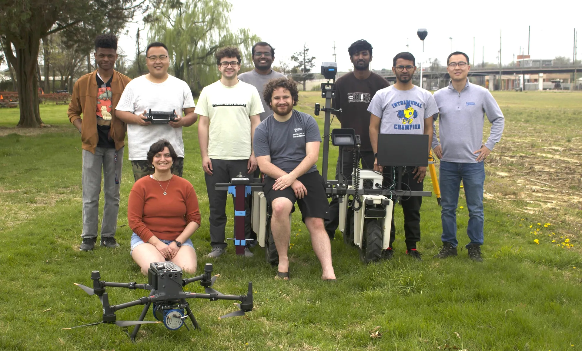 Seven guys stand around a robot, one holds a controller. A guy sits on the robot, while a woman sits on the grass behind the drone and beside the robot.