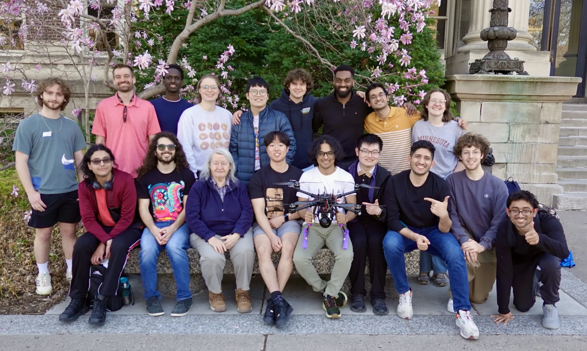 Eight people sit on a bench, one holding a drone, and one guy crouching at the end, while 9 people stand behind the bench.e