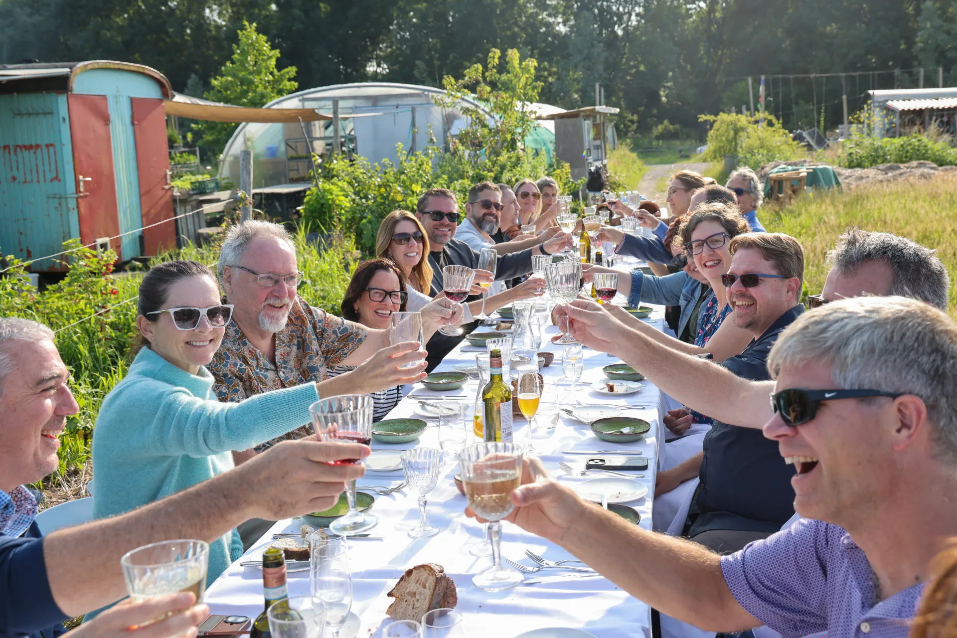 Large group of people seated at a long table, outdoors, raising their wine glasses in a toast
