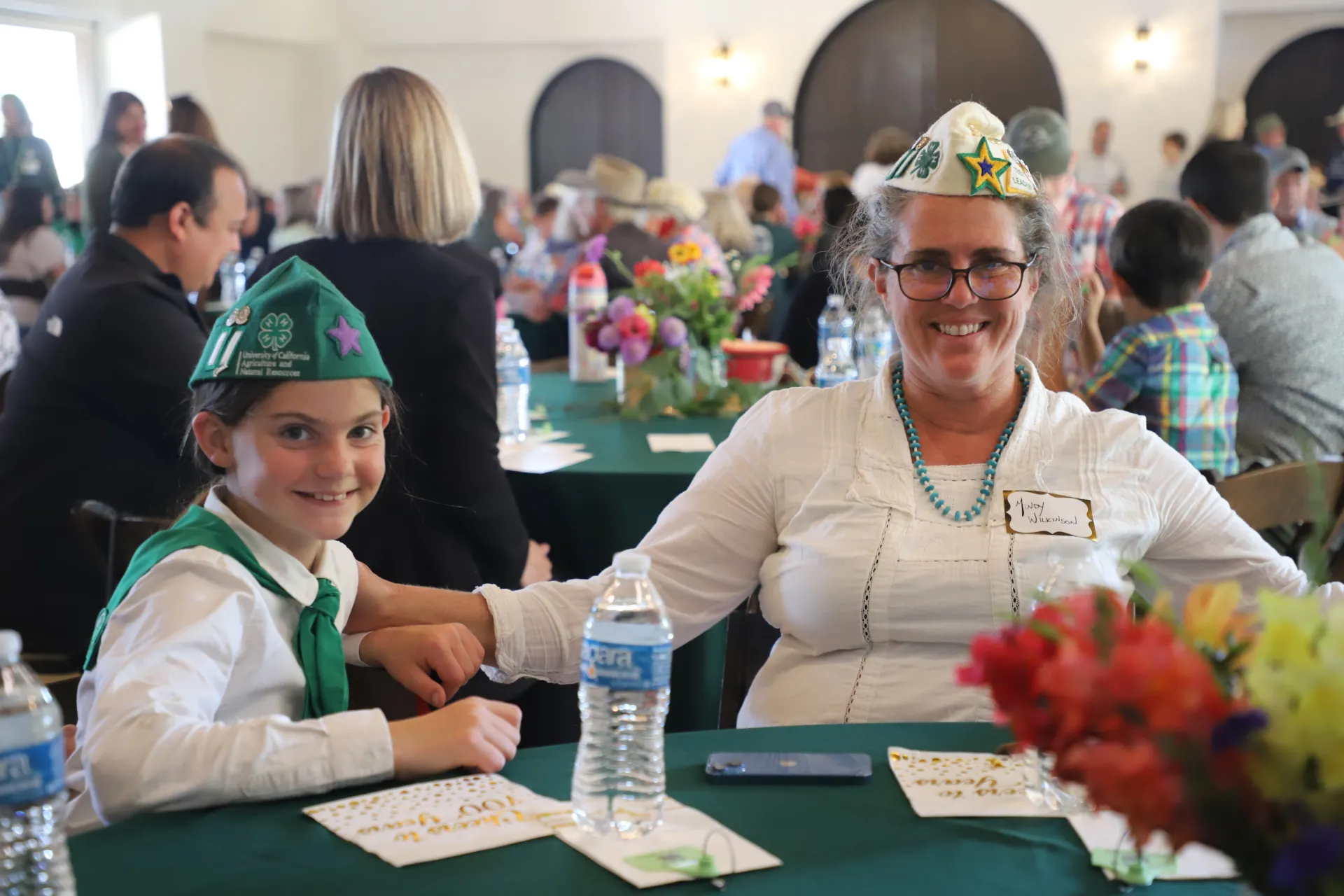 A girl and her mom, both in 4-H uniforms, sit at a table and smile at the camera.