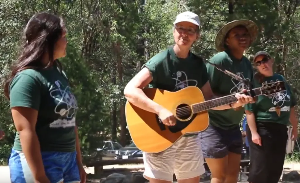Marianne strums guitar surrounded by 3 other women wearing matching dark green t-shirts