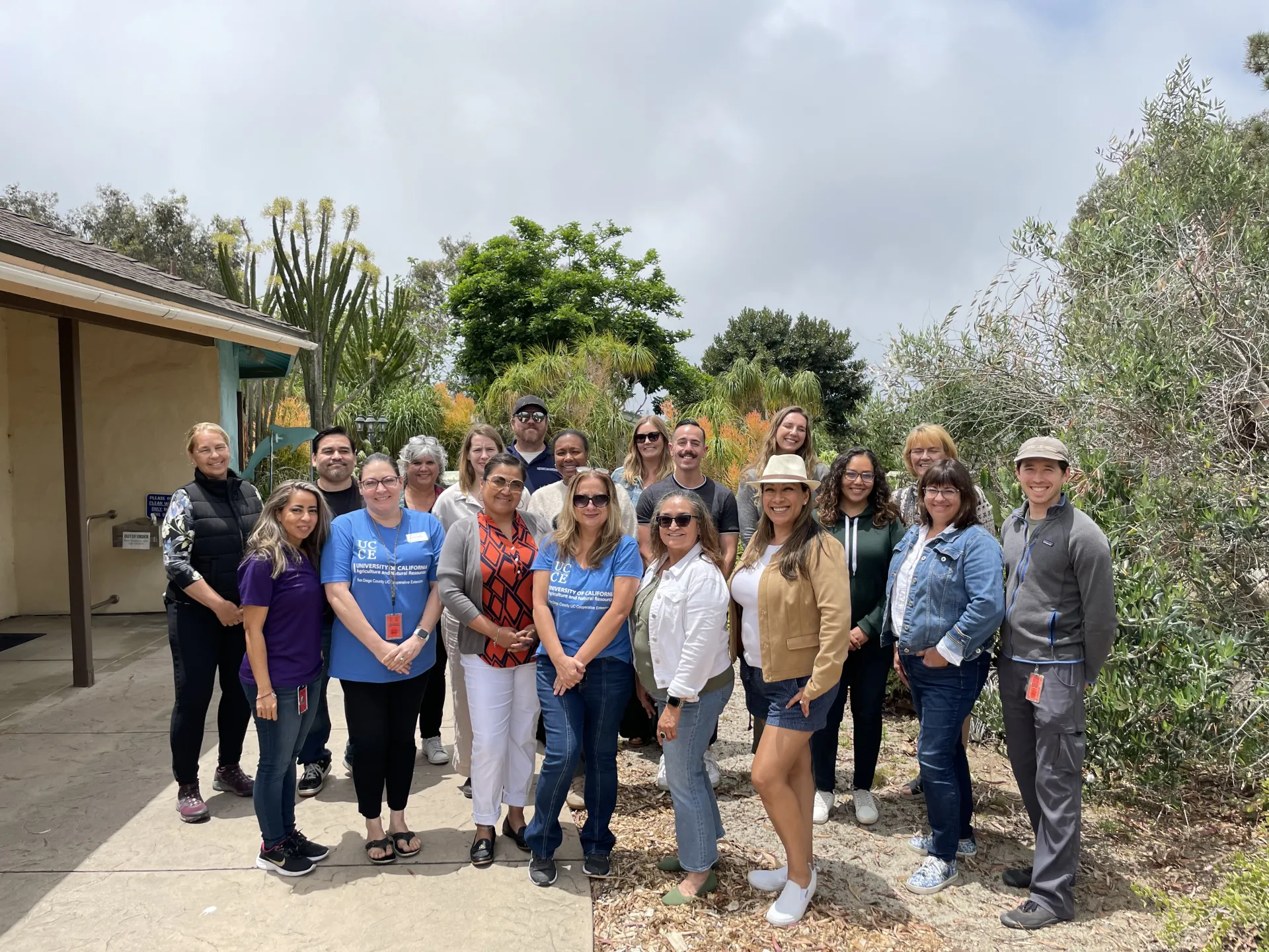 Nineteen people stand outdoors posing for a photo