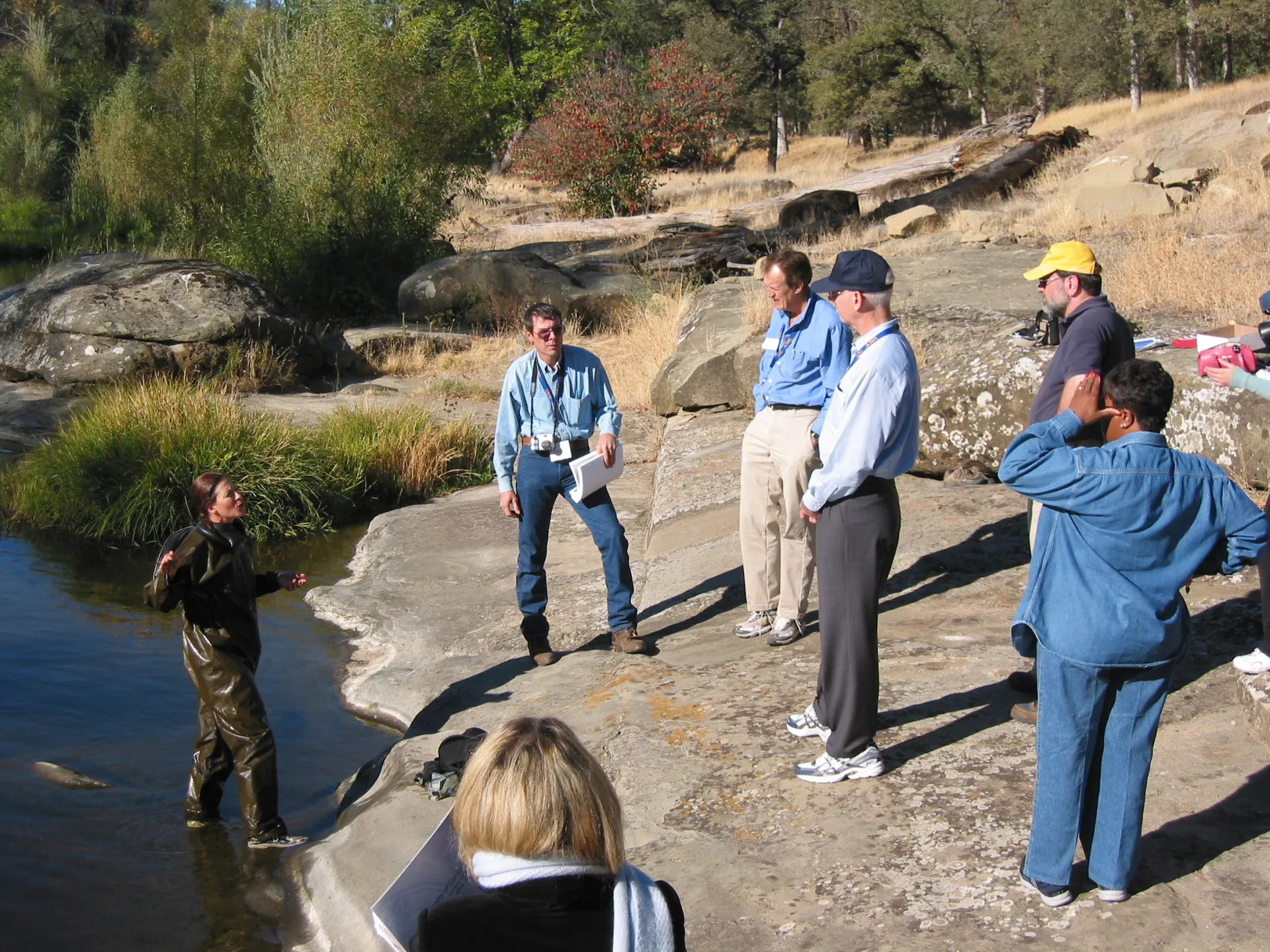 Lisa Thompson walks out of stream wearing a wet suit to talk to 6 people on the riverbank