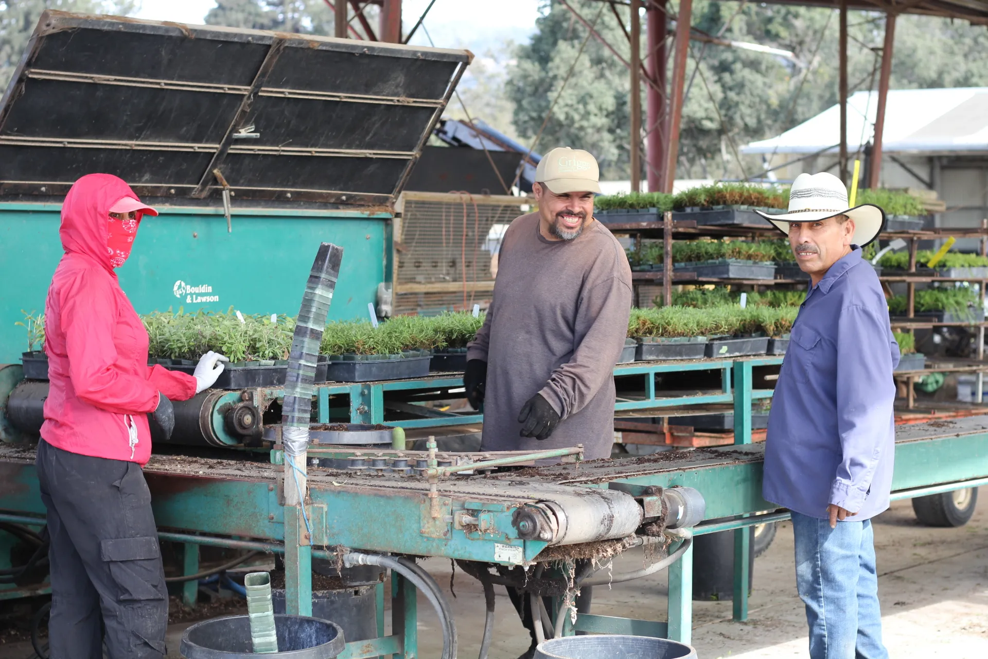 Three people stand in front of nursery machinery,