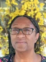 Michele J has dark-rimmed glasses and a dark blouse and is surrounded by yellow foliage.