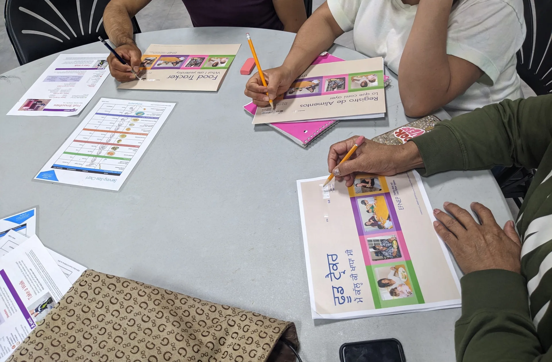 Three people sitting at the same table writing on worksheets that depict three different languages