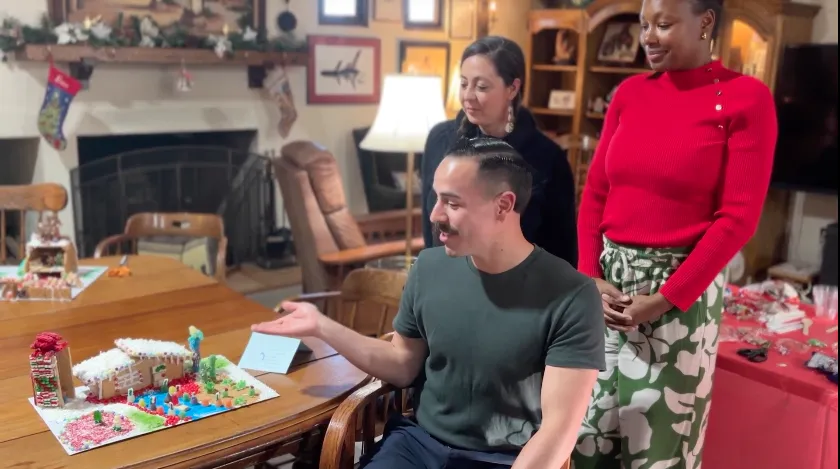Three people look at a gingerbread house surrounded by colorful candy landscaping