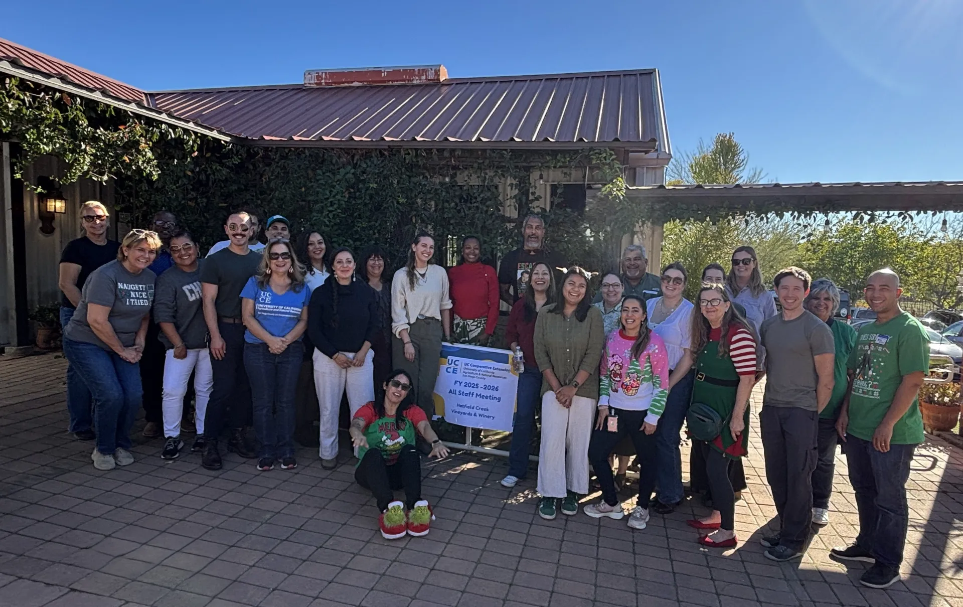 A group of 27 people pose outside around a sign that says "FY 2025-26 All Staff Meeting Hatfield Creek Vineyards & Winery 
