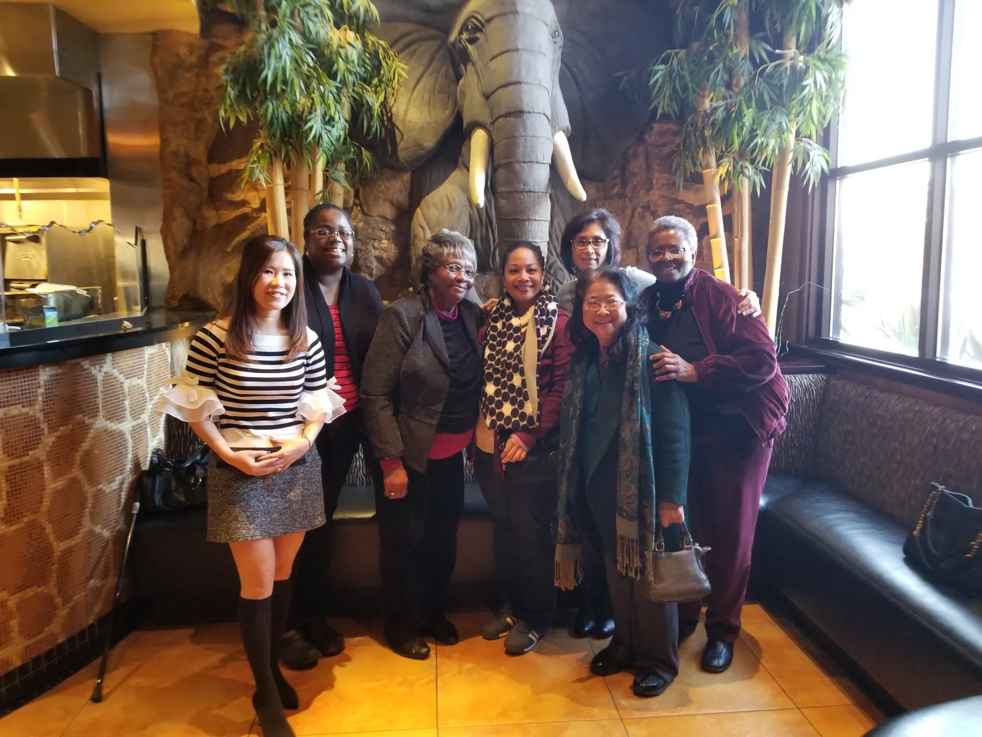 Seven women stand in front of an elephant statue and 2 trees