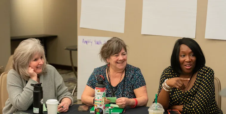 Three women, two white and one African-American, smile at a conference table.