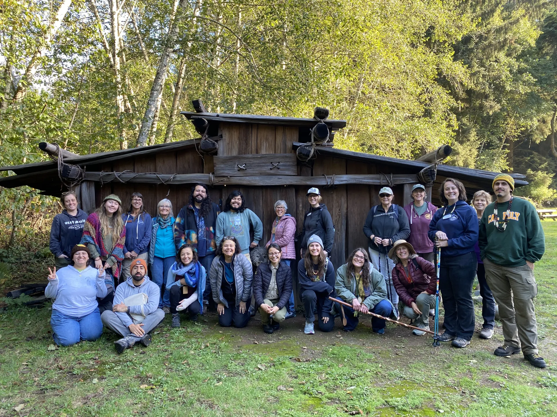 A group of men and women stand in front of a Yurok plank house, surrounded by the woods of a state park