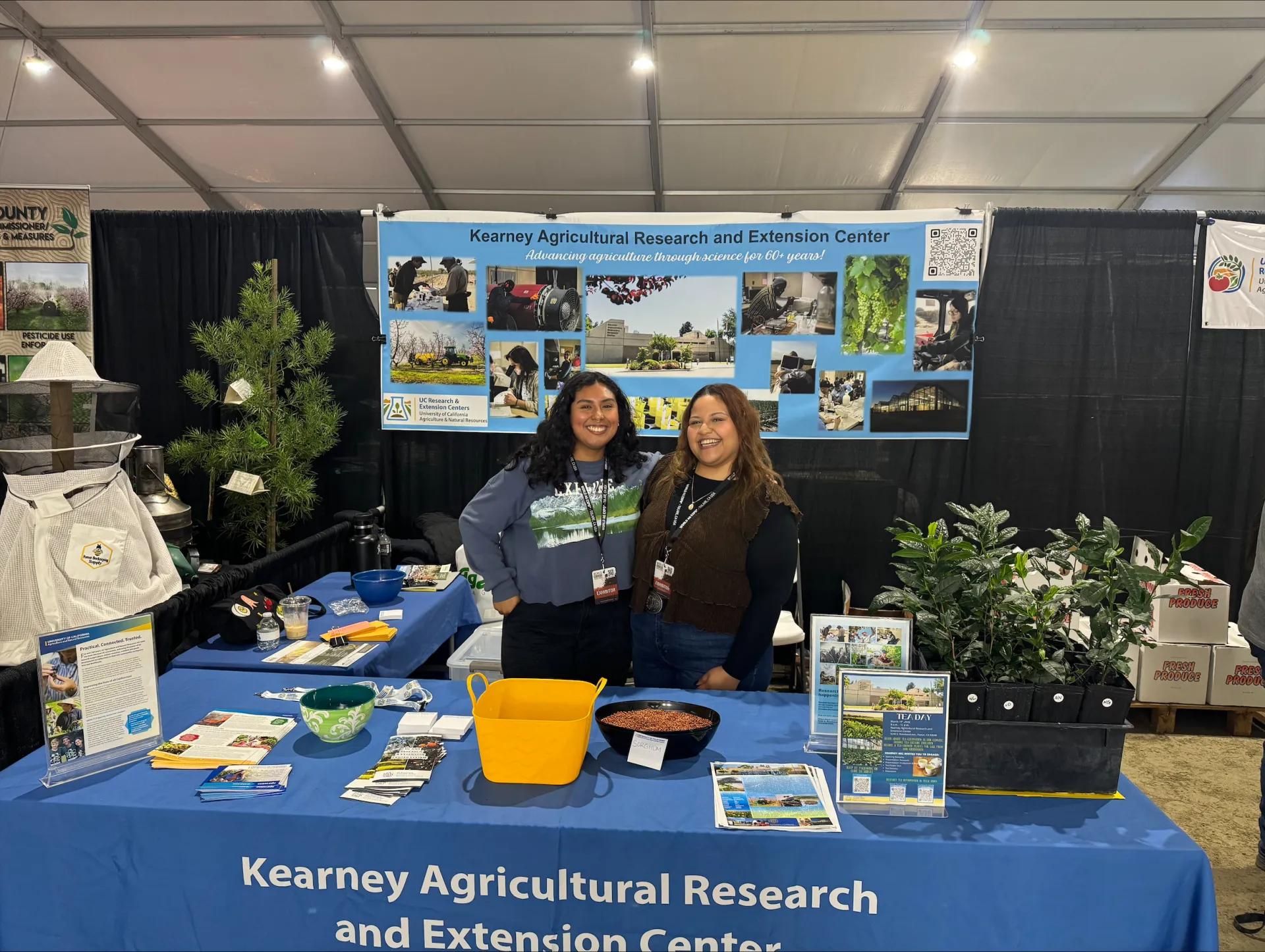 Nancy and Valeria stand between a banner and table, both labeled "Kearney Agricultural Research and Extension Center." The table displays tea plans and many paper handouts