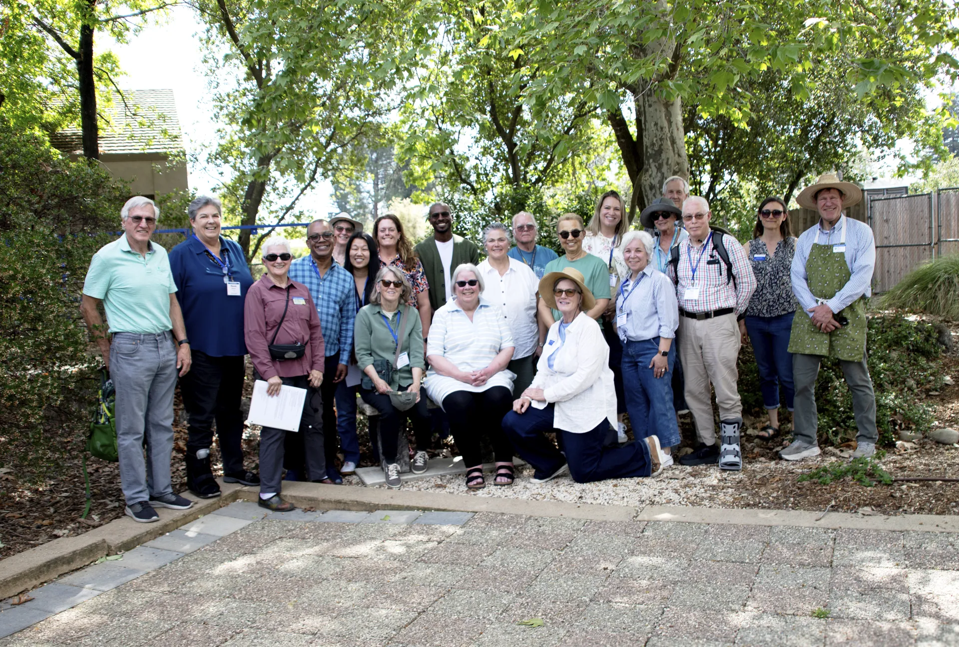 A group of 21 people pose in a garden under a leafy green tree