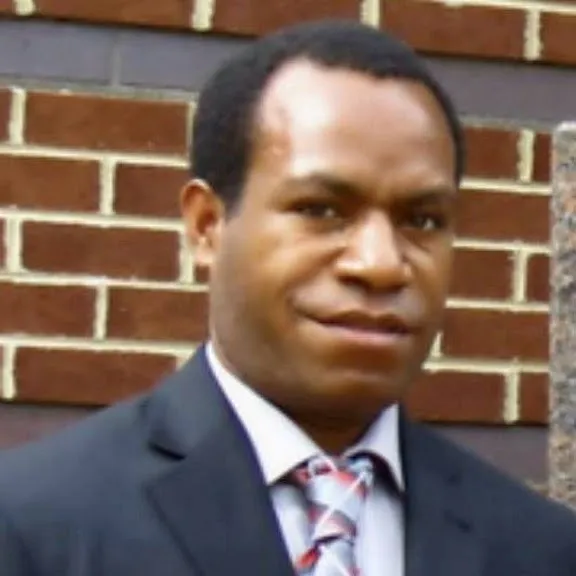 Close-up portrait of Philip Waisen smiling, wearing a navy blazer and tie, standing outside against a brick wall