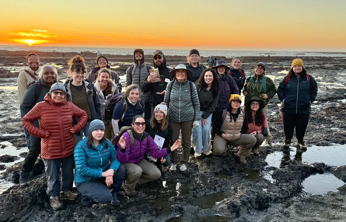 A group of about 20 participants in a UC California Naturalist course pose for a photo among the tidepools at Duxbury Reef as the sun sets behind them