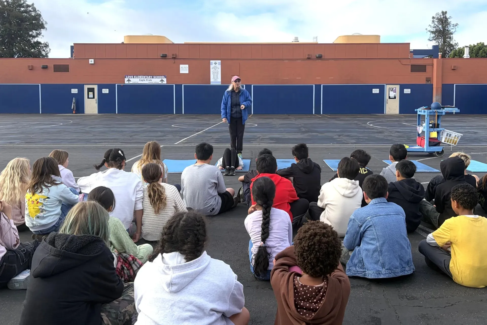 Una maestra de educacion fisica se dirige aun grupo de estudiantes sentados en el suelo.
