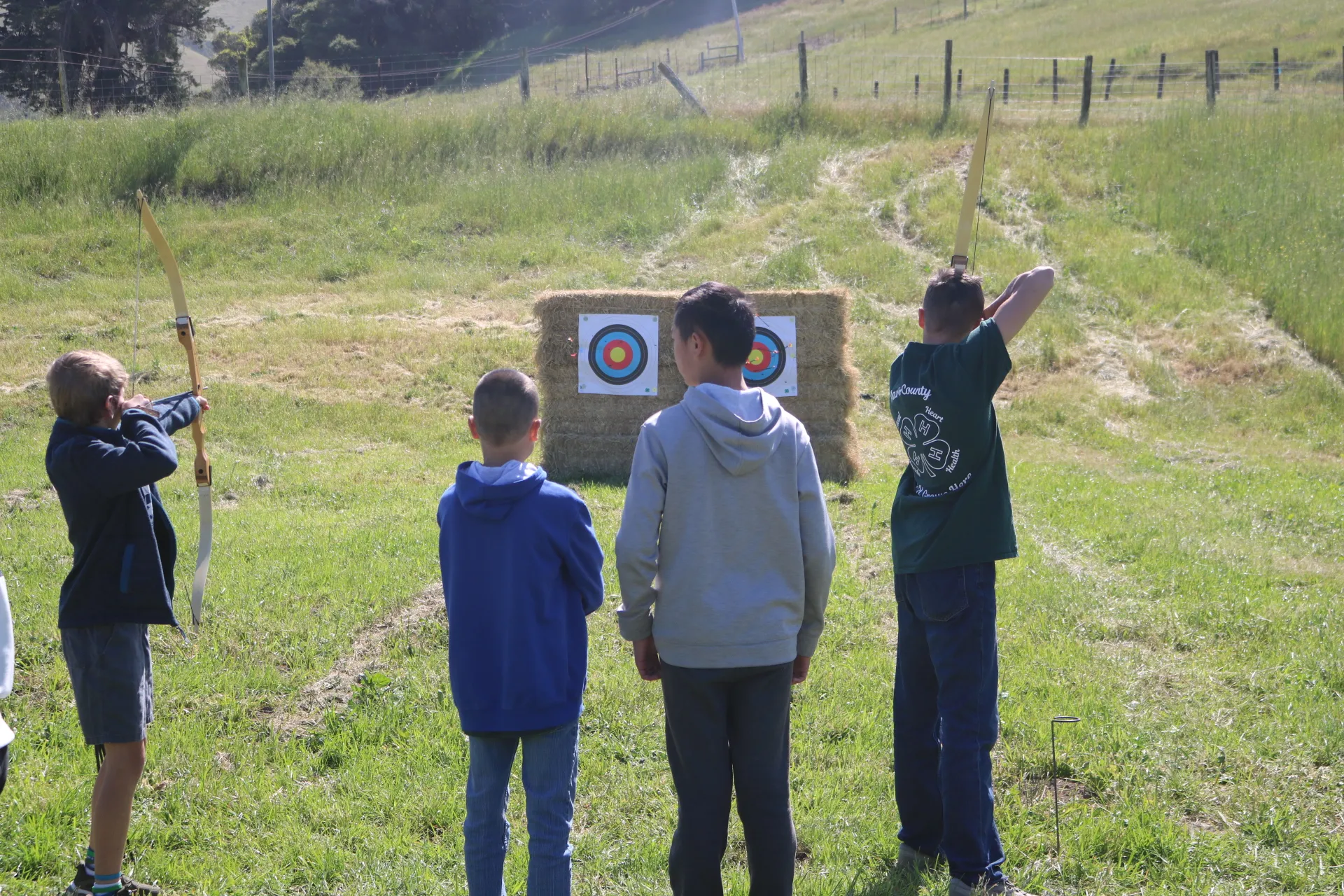 4 youth members in the frame. Two youth have a bow and arrow and two are standing behind as spectators. In the background are two archery targets on hay bales