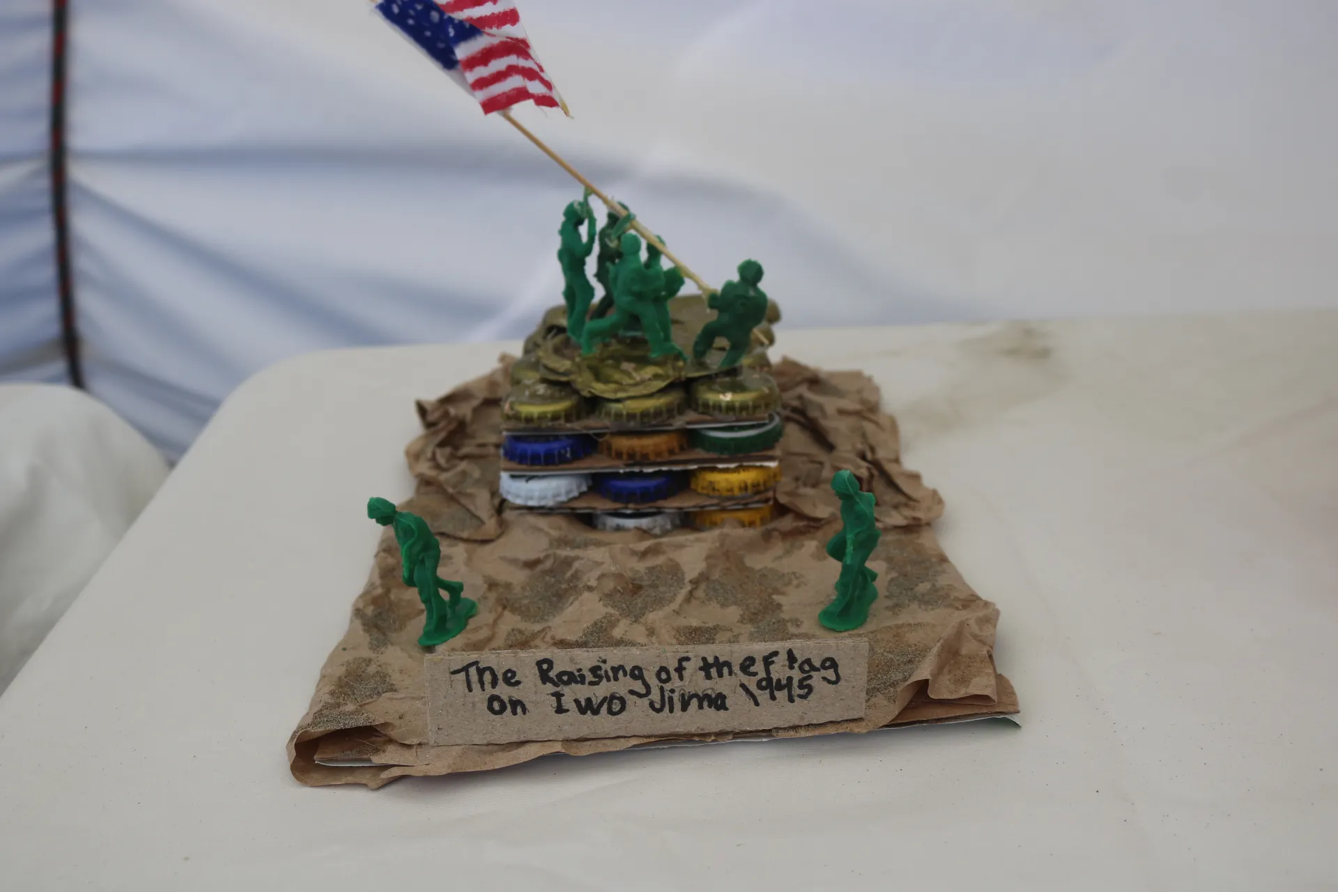 A recycled art piece diorama recreates the iconic raising of the American flag on Iwo Jima in 1945. Small green toy soldiers raise a miniature U.S. flag atop a hill constructed from stacked bottle caps and cardboard, with crumpled brown paper and sand representing the rocky landscape. Additional toy soldiers stand at the base of the hill, and a handwritten label on the front reads, "The Raising of the Flag on Iwo Jima 1945."