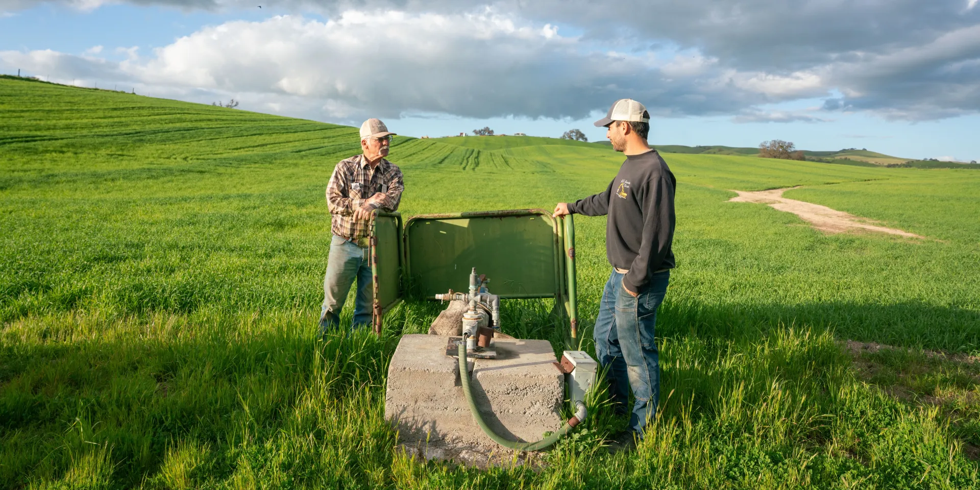 Two farmers stand in a vast grazing field, looking at a well pump