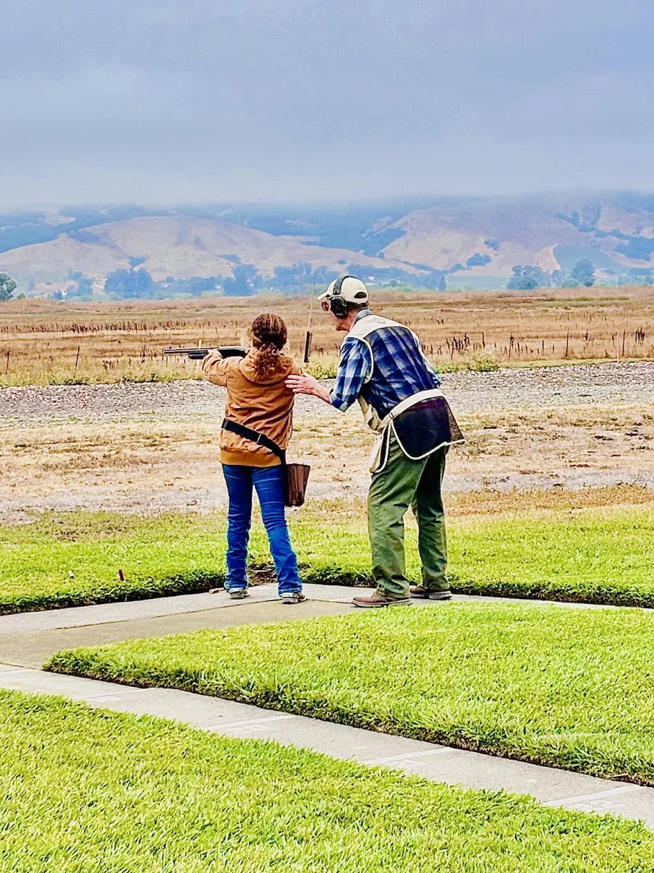 a girl holding a shotgun with an adult male instructor next to her looking in the distance. Green grass with cement pathways are seen on the ground with a mountain landscape in the background. 
