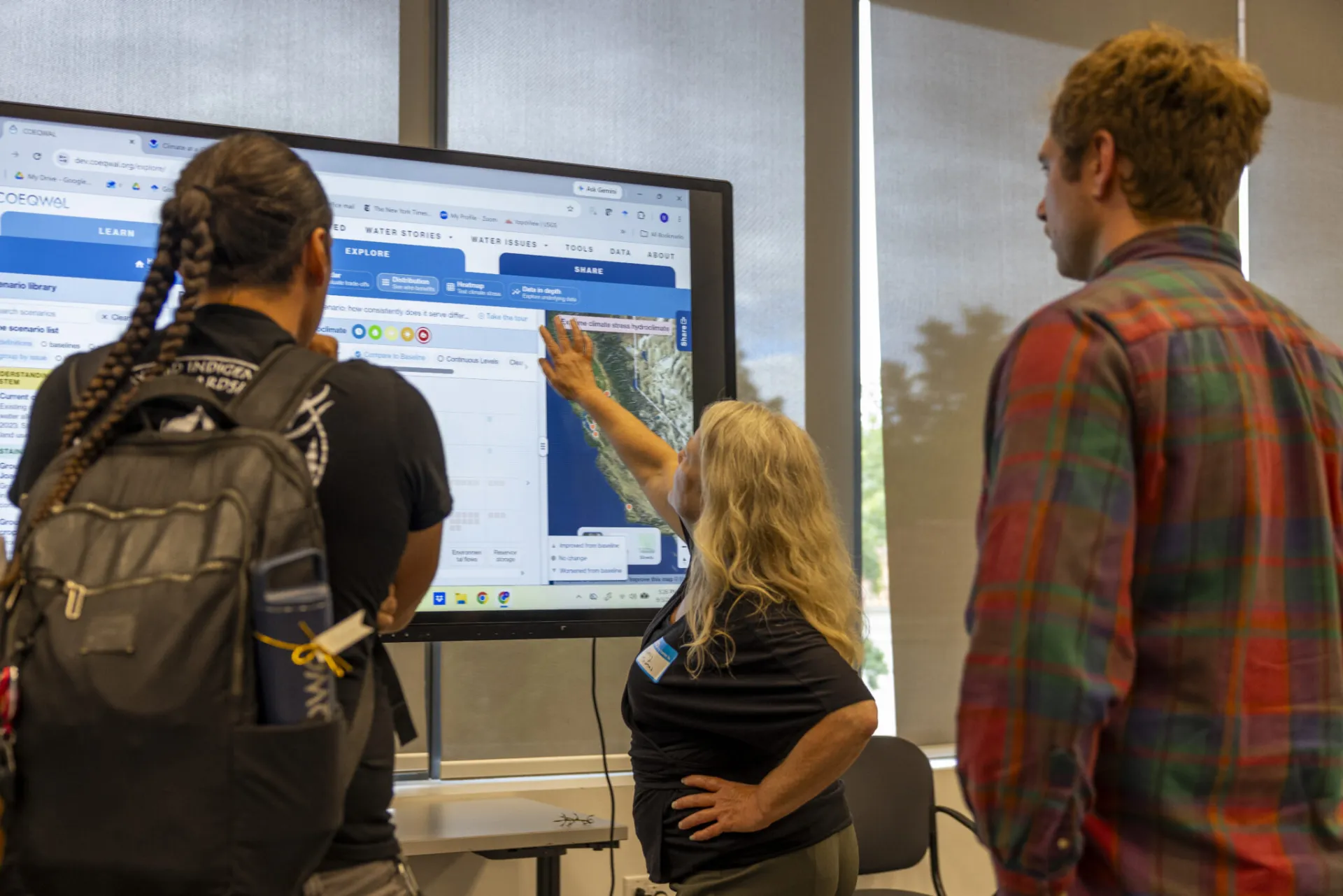 A woman points to a map on a large computer screen as two people look on 