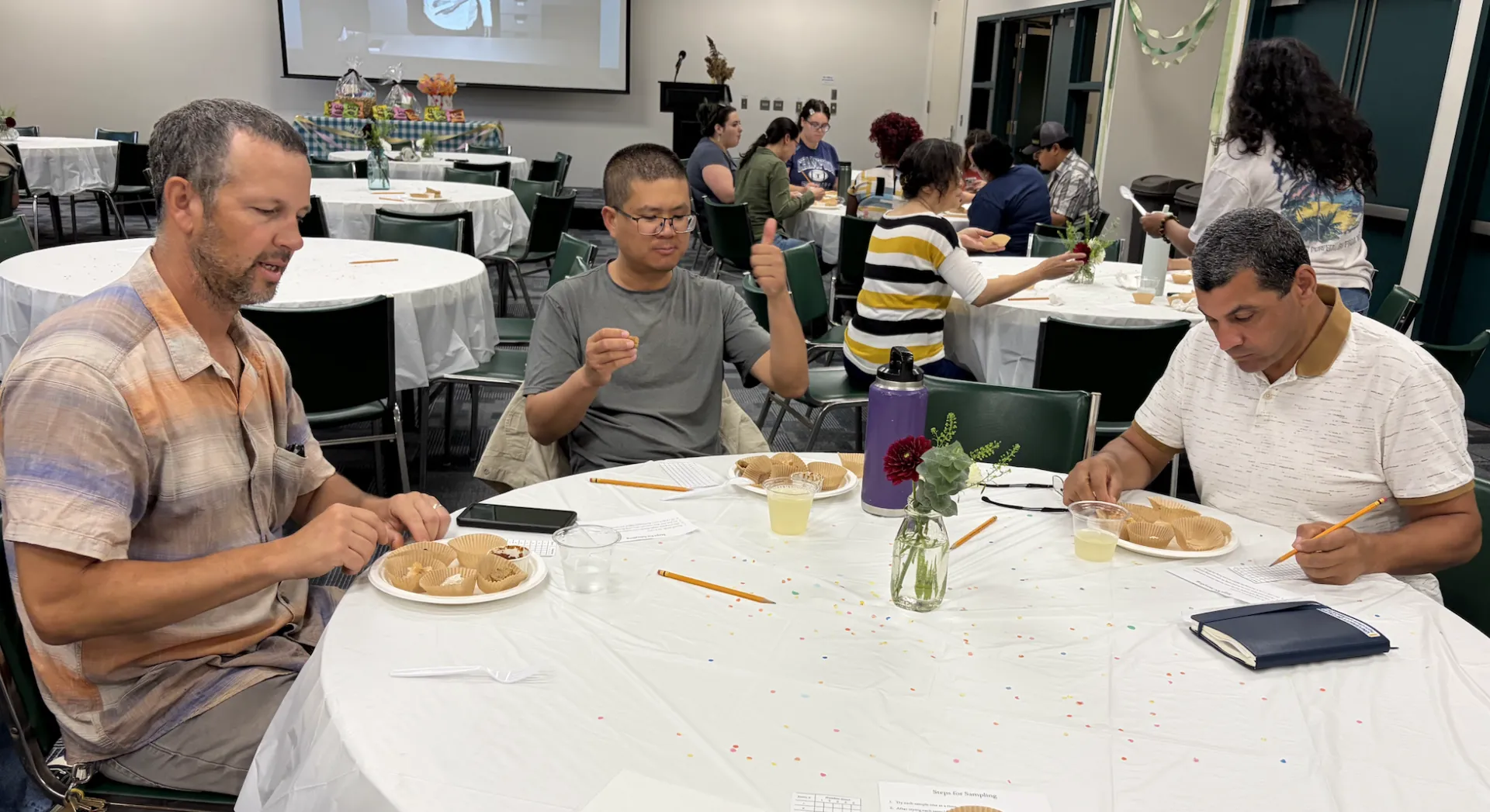 People seated at round tables with white tablecloths taste food samples. George gives a thumbs up