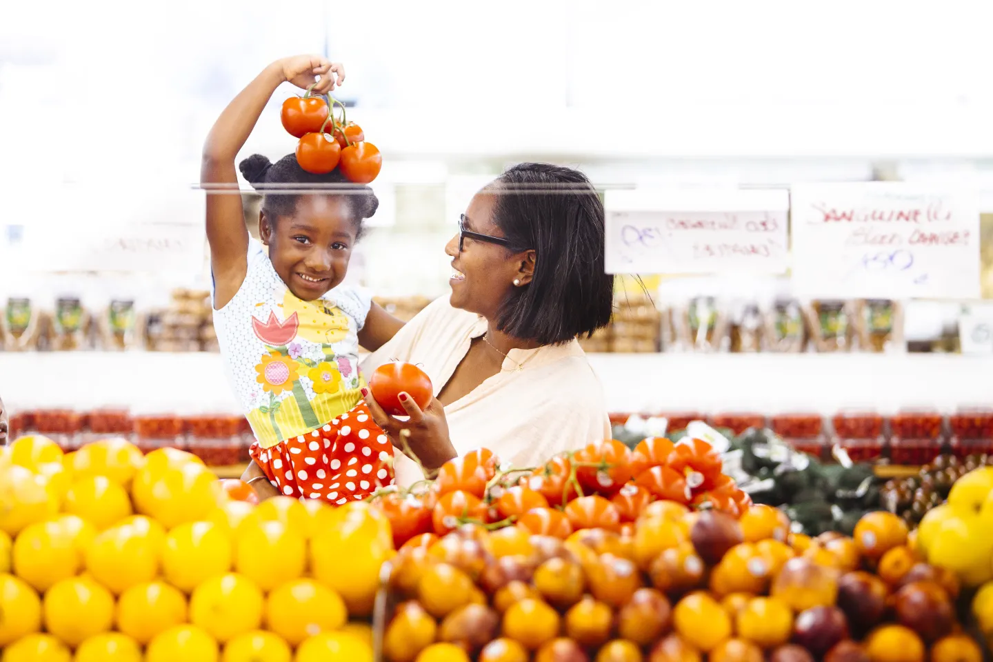 A child holds a cluster of tomatoes over her head as she smiles at a grocery store