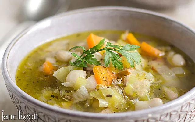 Bowl of vegetable soup with carrots, leeks, white beans, and a garnish of parsley, served in a textured ceramic bowl.