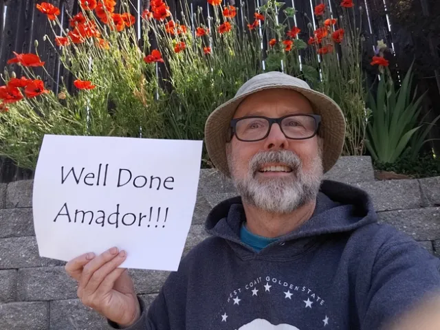 Photo of UC Master Gardener volunteer Ed Bass wearing a gardening hat and glasses, holding up a sign in his gardener reading