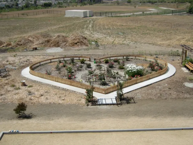 A heart-shaped fence surrounds a garden bed. At the tip of the heart, an arbor showcases pink climbing roses. Inside the heart-shaped fence are 20 rose bushes in colors such as white, red, purple, and violet.