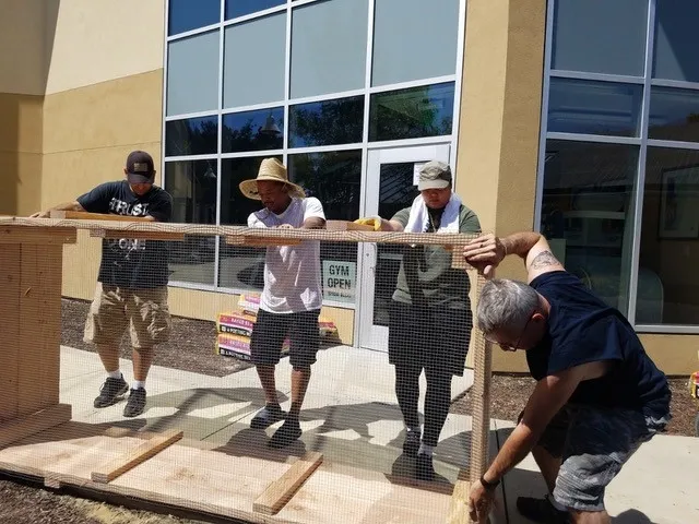Four people building a raised garden bed, the garden bed is on its side with a wire mesh being attached to the bottom to prevent pests.