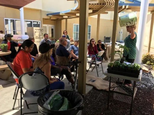 A group of 11 participants sitting under a trellis as a speaker in the front of the group holds up a bucket, standing next to a table of young tomato plants.