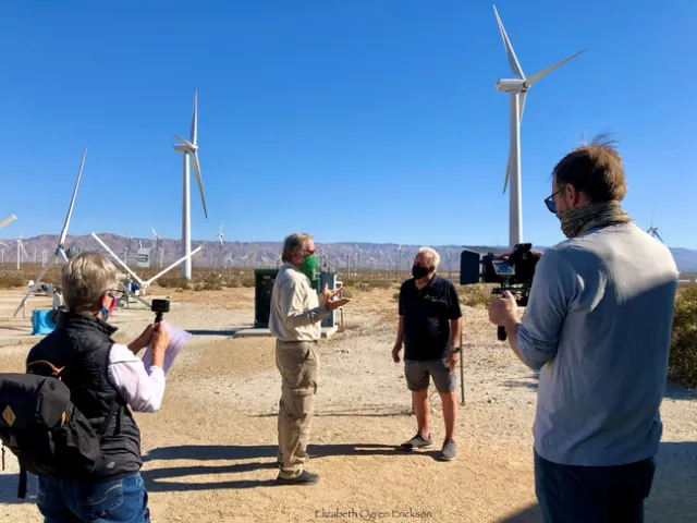 With everyone wearing face masks, the four people stand on dusty soil among dozens of three-bladed windmills, under a cloudless blue sky.