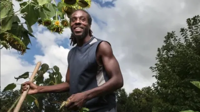 Sacramento farmer Chanowk Yisrael, shown from the waist up holding a shovel handle, stands among crops.