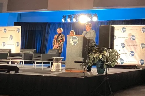 Three women in suits stand on stage. Glenda Humiston speaks from behind a lecturn.