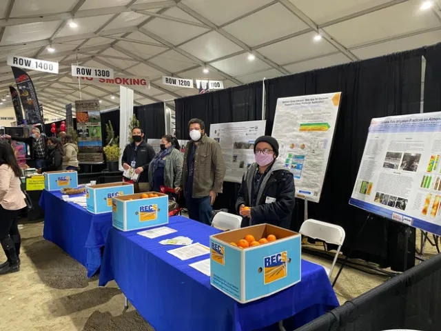 The four people wear face masks and stand behind a table with boxes of mandarins. Behind them are research posters on easels.