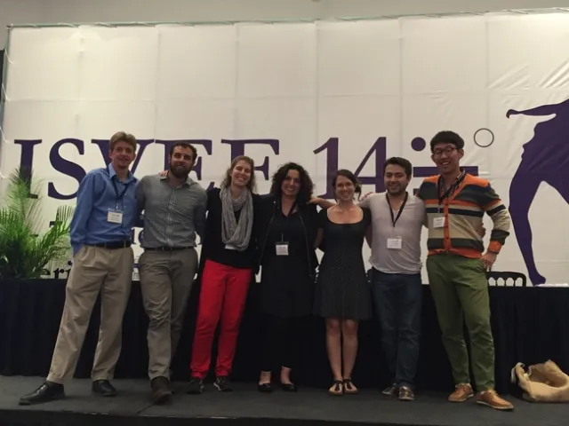 Seven people stand in a row in front of a conference banner.
